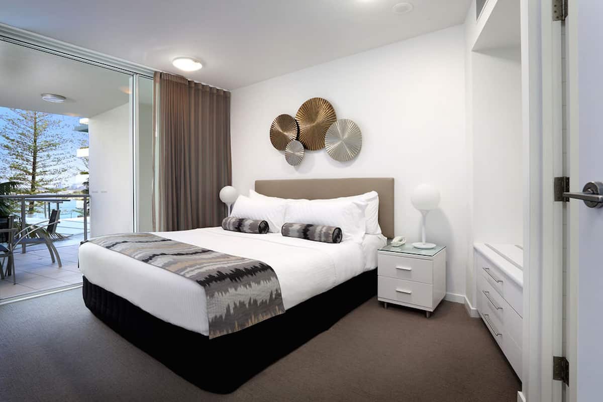 A modern hotel room with a large bed, a bedside table, a nightstand, and a view of the outdoors.