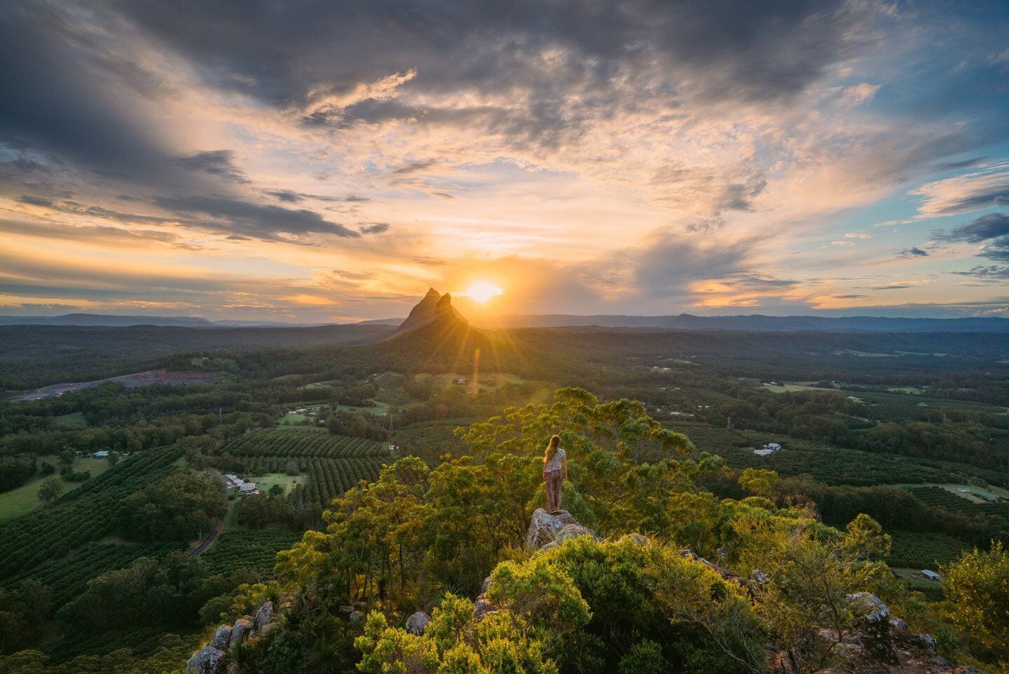 A person standing on a hill overlooking a valley with a mountain in the distance and the sun setting behind it.