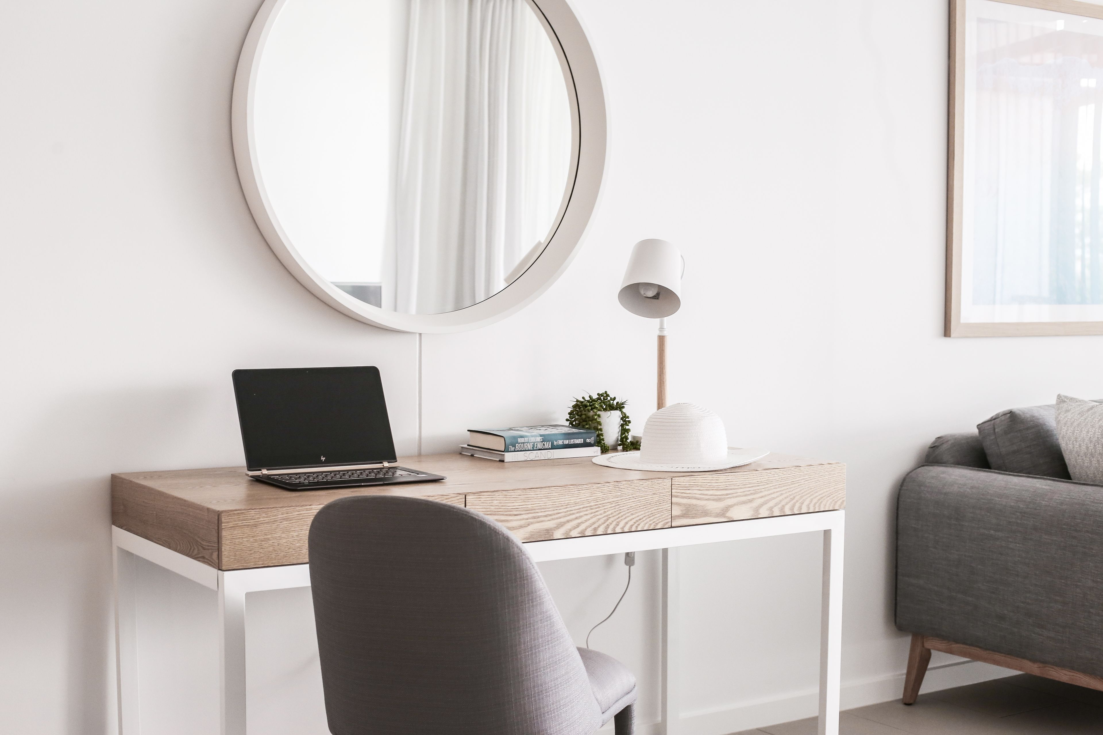 A modern workspace with a laptop, books, a lamp, and a round mirror on a wooden desk.