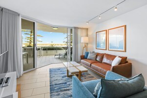 Una sala de estar moderna con un sofá de cuero marrón, un sillón azul y una mesa de centro. Hay una puerta corrediza de vidrio que da a un patio exterior con vistas a árboles y un cielo despejado.