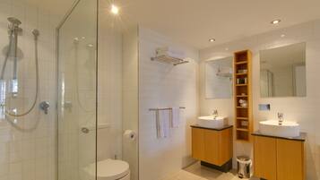A modern bathroom with a glass-enclosed shower, a toilet, two sinks, and a vanity with storage shelves.