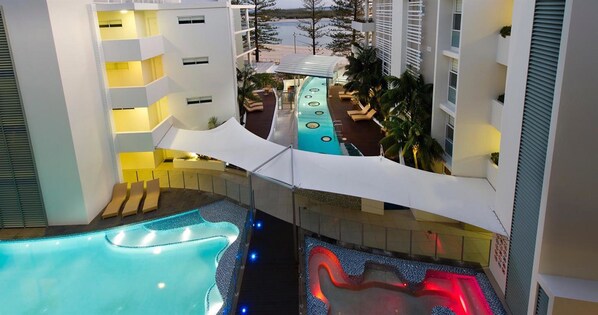 Un hotel de varios niveles con zona de piscina, que incluye un jacuzzi y sillones de descanso.
