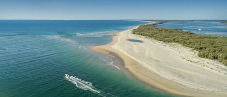 An aerial view of a coastal area with a sandy beach, clear blue waters, and a forested area.