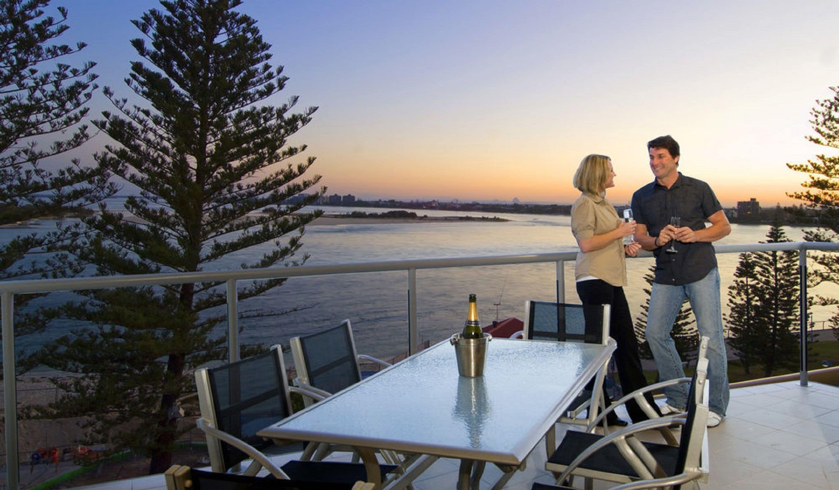 A couple standing on a balcony overlooking a river with a sunset view, a table with chairs, and a bottle of champagne.