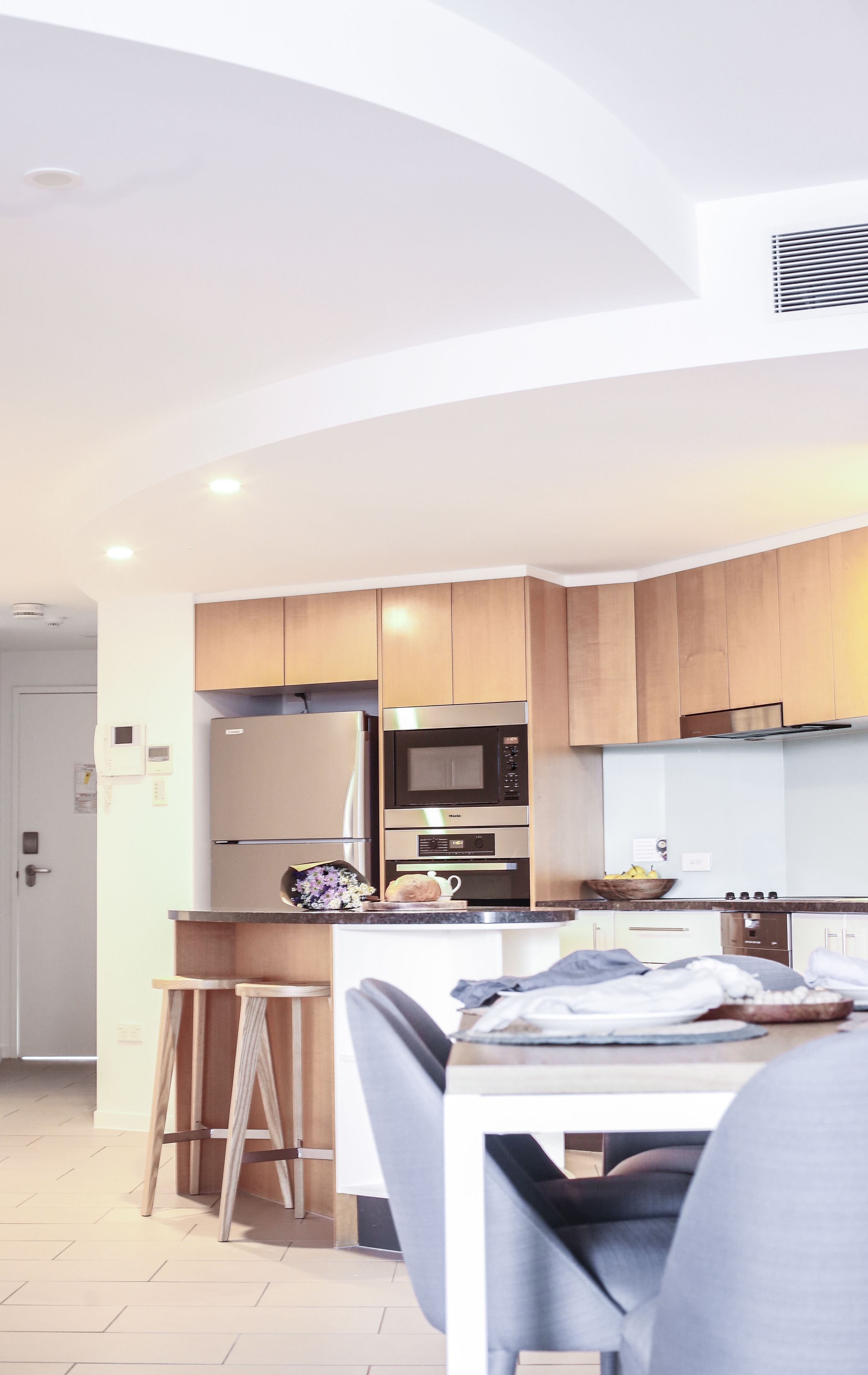 A modern kitchen with wooden cabinets, a stainless steel refrigerator, a microwave, and a built-in oven. There is a small island with stools and a table with a bowl of fruit.