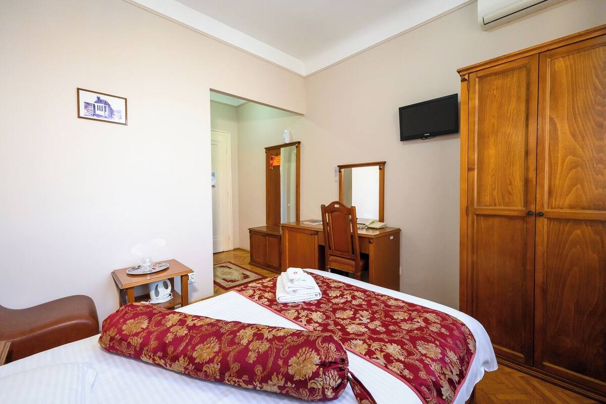Ein Hotelzimmer mit Bett, einem Holzschrank, einem Schreibtisch mit Stuhl, einem Spiegel und einem Fernseher an der Wand.