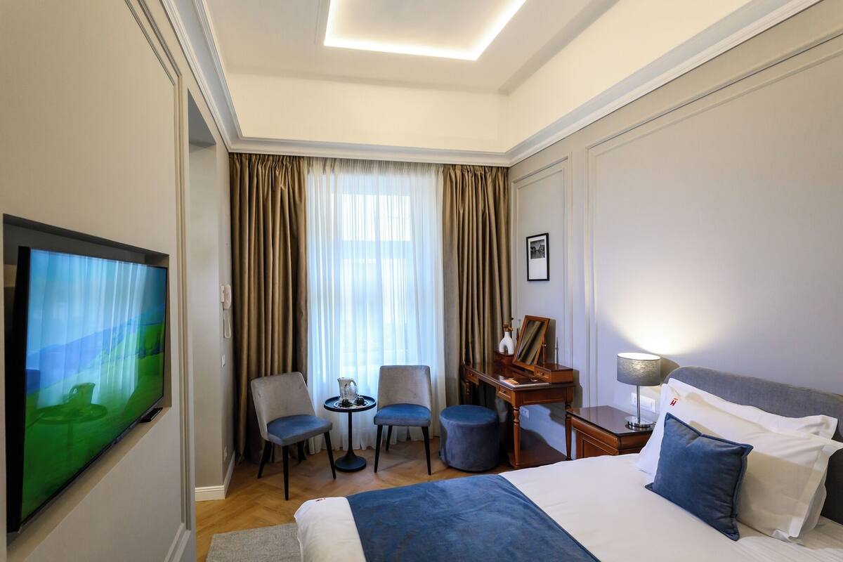 Ein Hotelzimmer mit einem großen Bett, einem Fernseher, einem Schreibtisch mit Lampe, zwei Stühlen, einem kleinen Tisch mit einem Krug und einem Fenster mit Vorhängen.