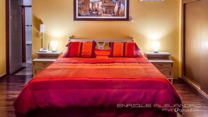 Quarto de hotel com uma cama grande, duas mesas de cabeceira com luminárias, um quadro na parede e uma porta à esquerda.