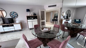 A modern dining area with a glass table surrounded by pink chairs, a white cabinet with decorative items, and a television in the background. - Riviera Eden Palace (Cannes)