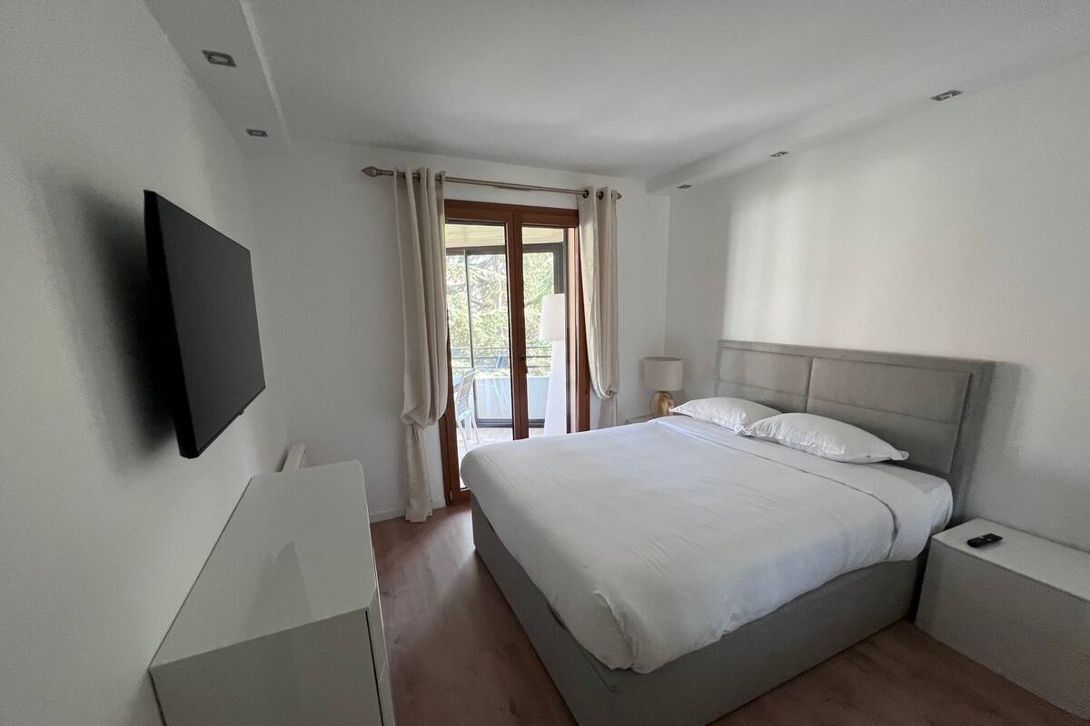 A modern bedroom with a large bed, bedside tables, a flat-screen TV, and a sliding glass door leading to a balcony.