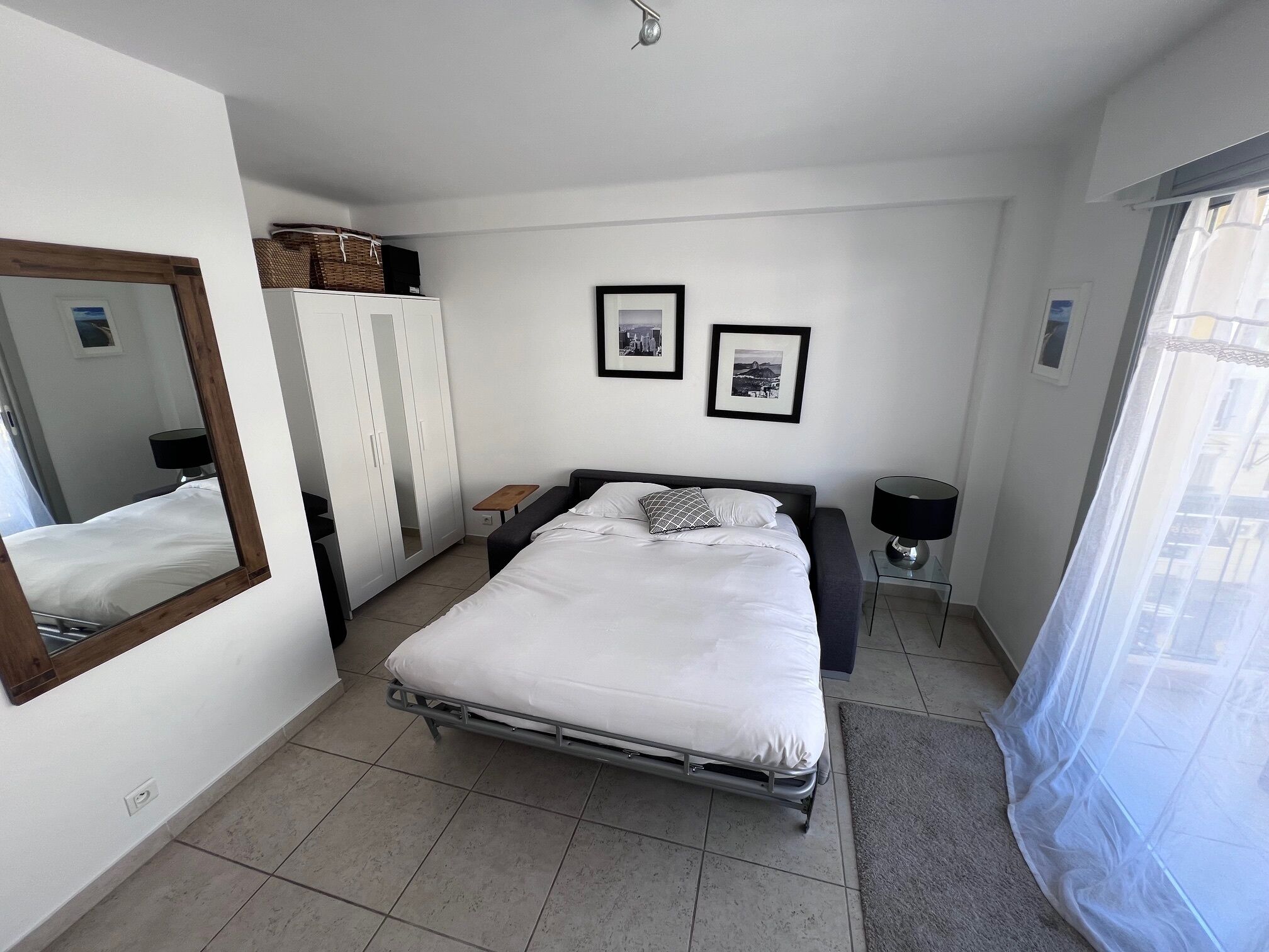 Une chambre à coucher comprenant un lit, une armoire, un miroir, deux tableaux encadrés, une table de chevet et une fenêtre avec un rideau léger.