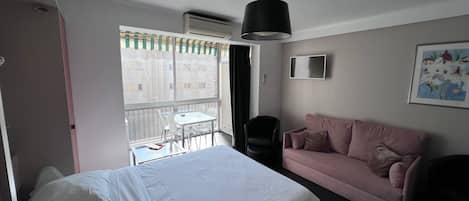 Une chambre d’hôtel avec un lit, un canapé rose, une petite table et une chaise. Il y a une fenêtre qui donne sur les bâtiments à l’extérieur.