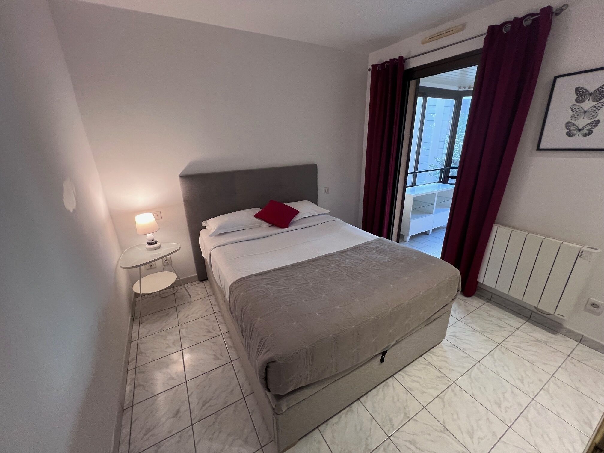 A bedroom with a bed, a nightstand, a lamp, a window with curtains, and a radiator.