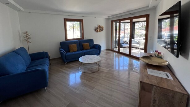 A modern living room with a blue sofa, a round coffee table, and a large window providing natural light. - Riviera Eden Palace (Cannes)