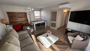 A modern living room with a sofa, coffee table, and a dining table with chairs. There is a TV mounted on the wall and a door leading to another room. - Riviera Eden Palace (Cannes)