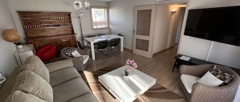 Ein modernes Wohnzimmer mit einer Couch, einem Couchtisch und einem Esstisch mit Stühlen. An der Wand ist ein Fernseher angebracht und eine Tür führt in einen anderen Raum.