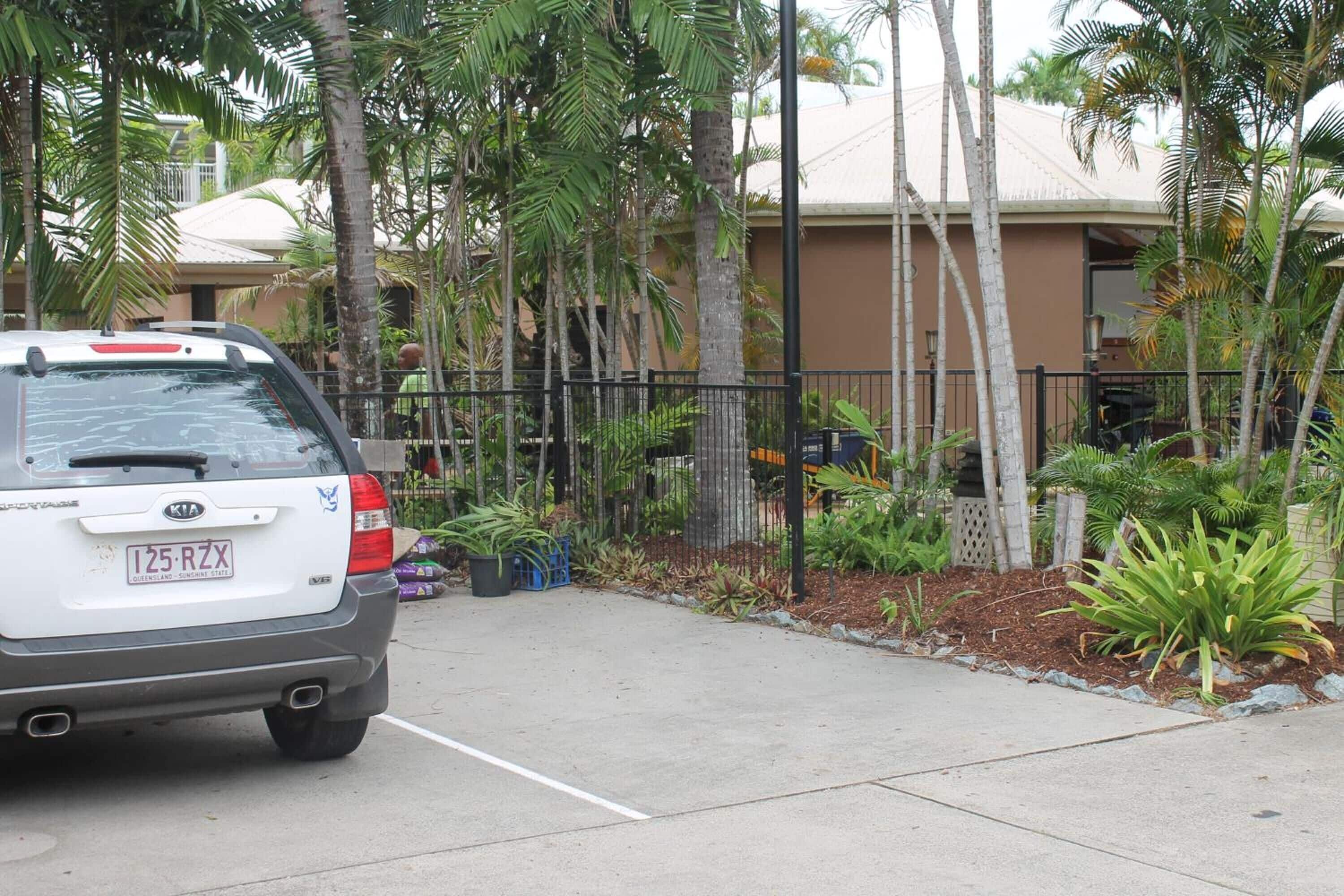 A white Kia SUV parked in a driveway with a concrete path leading to a house surrounded by palm trees and a black metal fence.