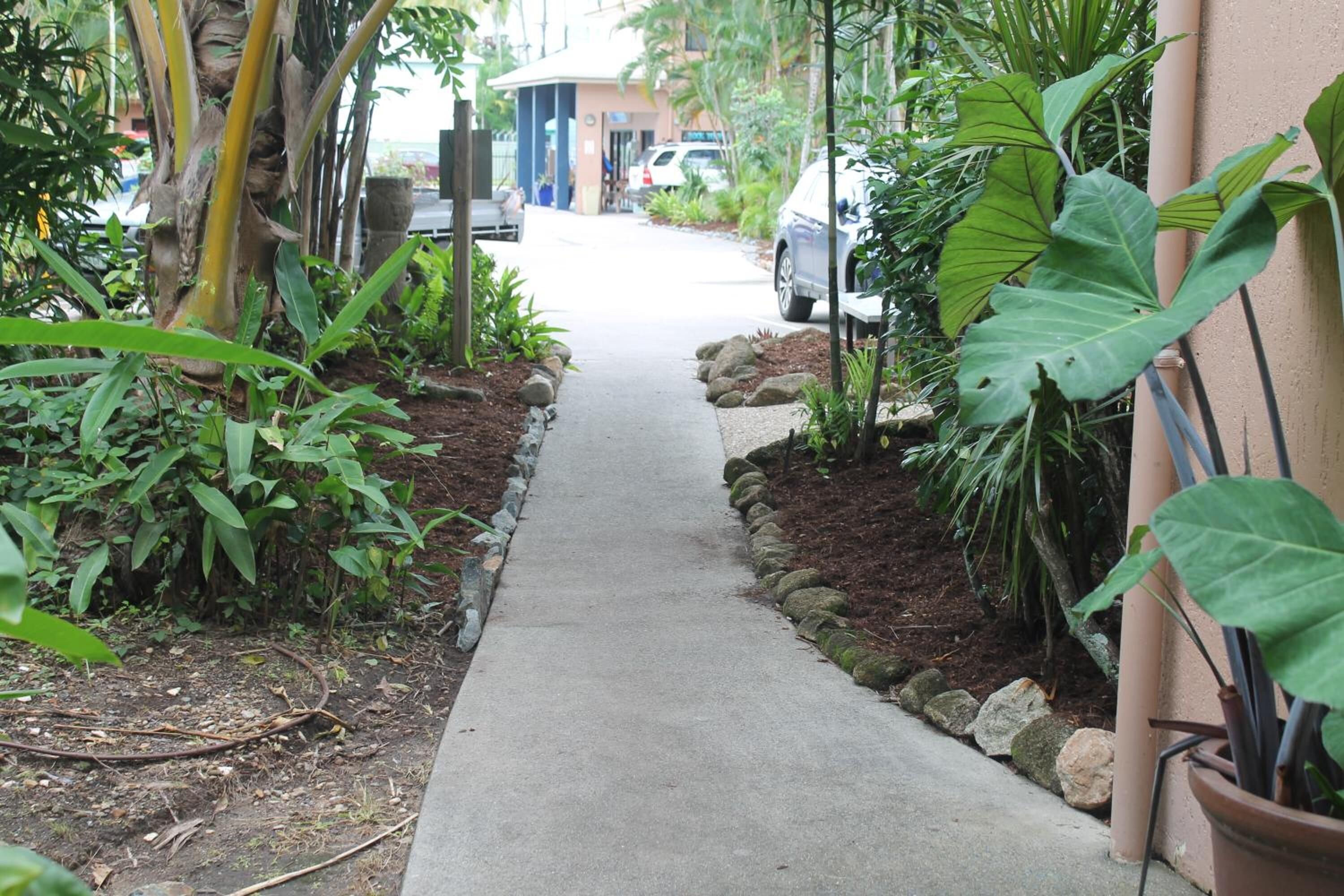 Un sendero de concreto bordeado de rocas y plantas, que conduce a un edificio de fachada color crema.
