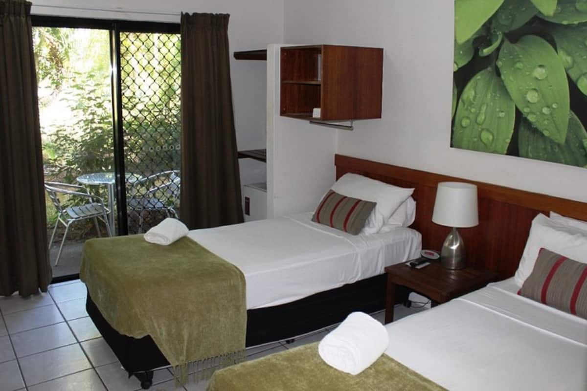 A hotel room with two beds, a wooden cabinet, a table with chairs, and a view of greenery through a window.