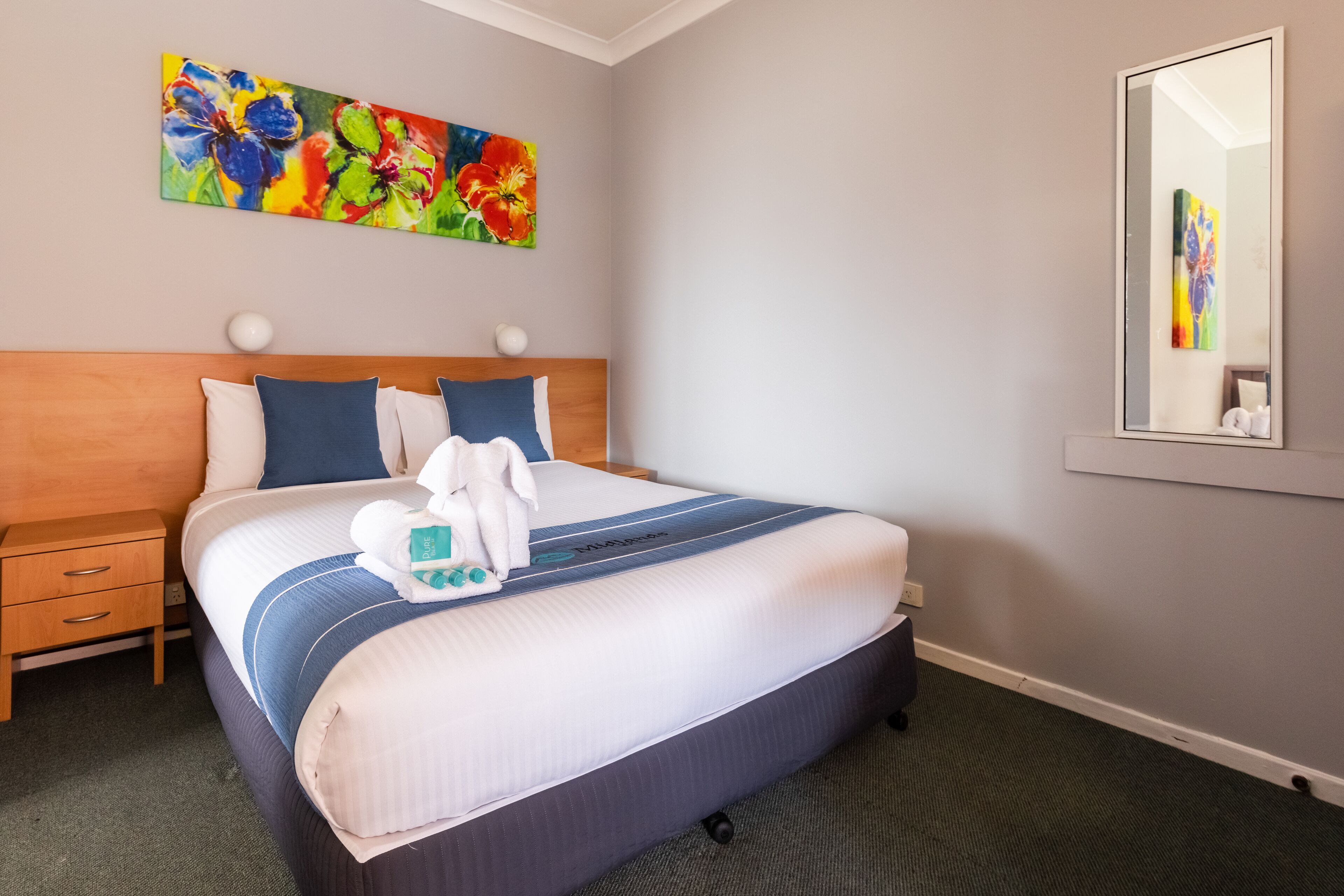 Una cama bien hecha con colcha azul y blanca a rayas, cabecero de madera y un colorido cuadro floral en la pared superior.