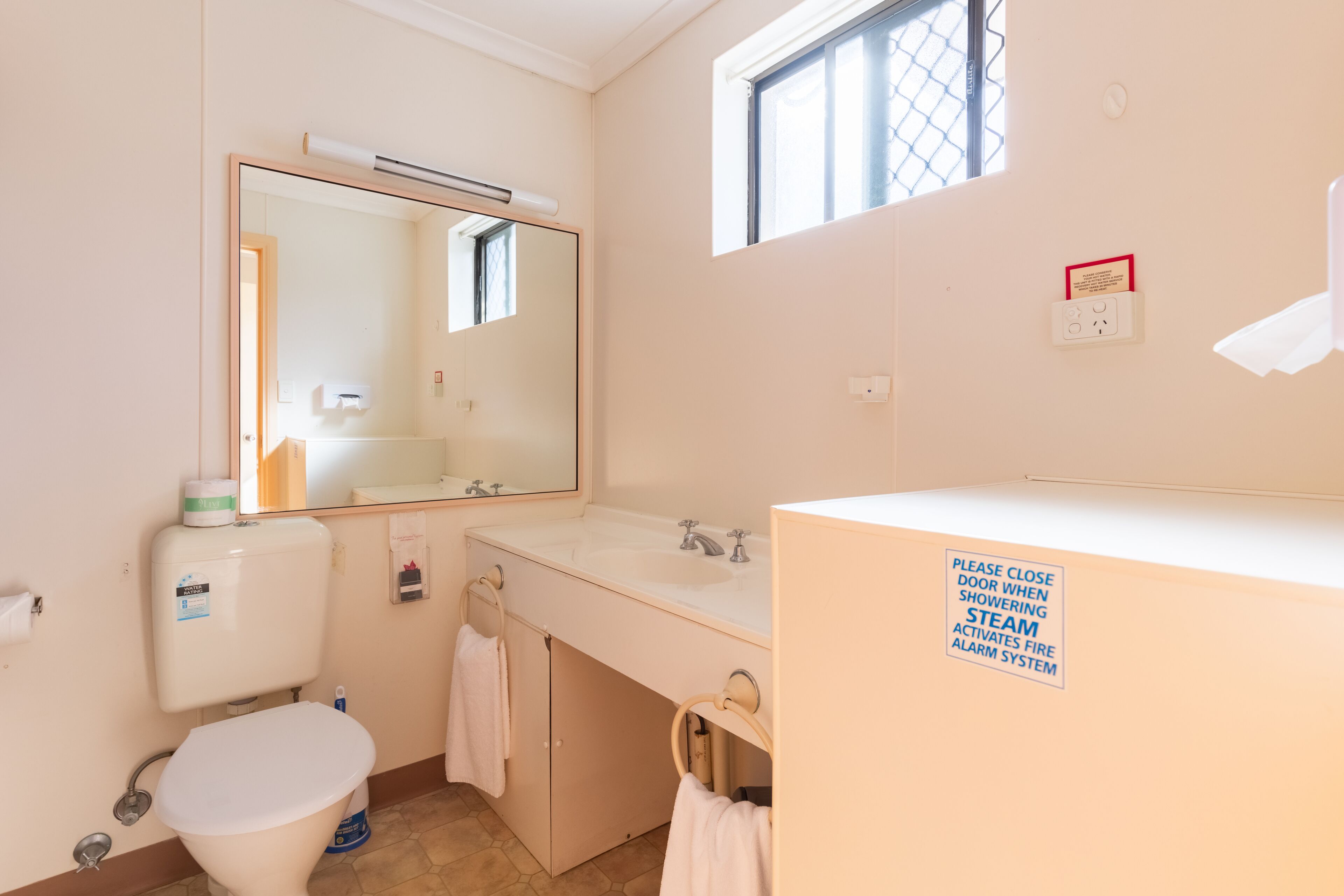A bathroom with a toilet, sink, and mirror. There is a sign instructing to close the door when the showering steam activates the fire alarm system.