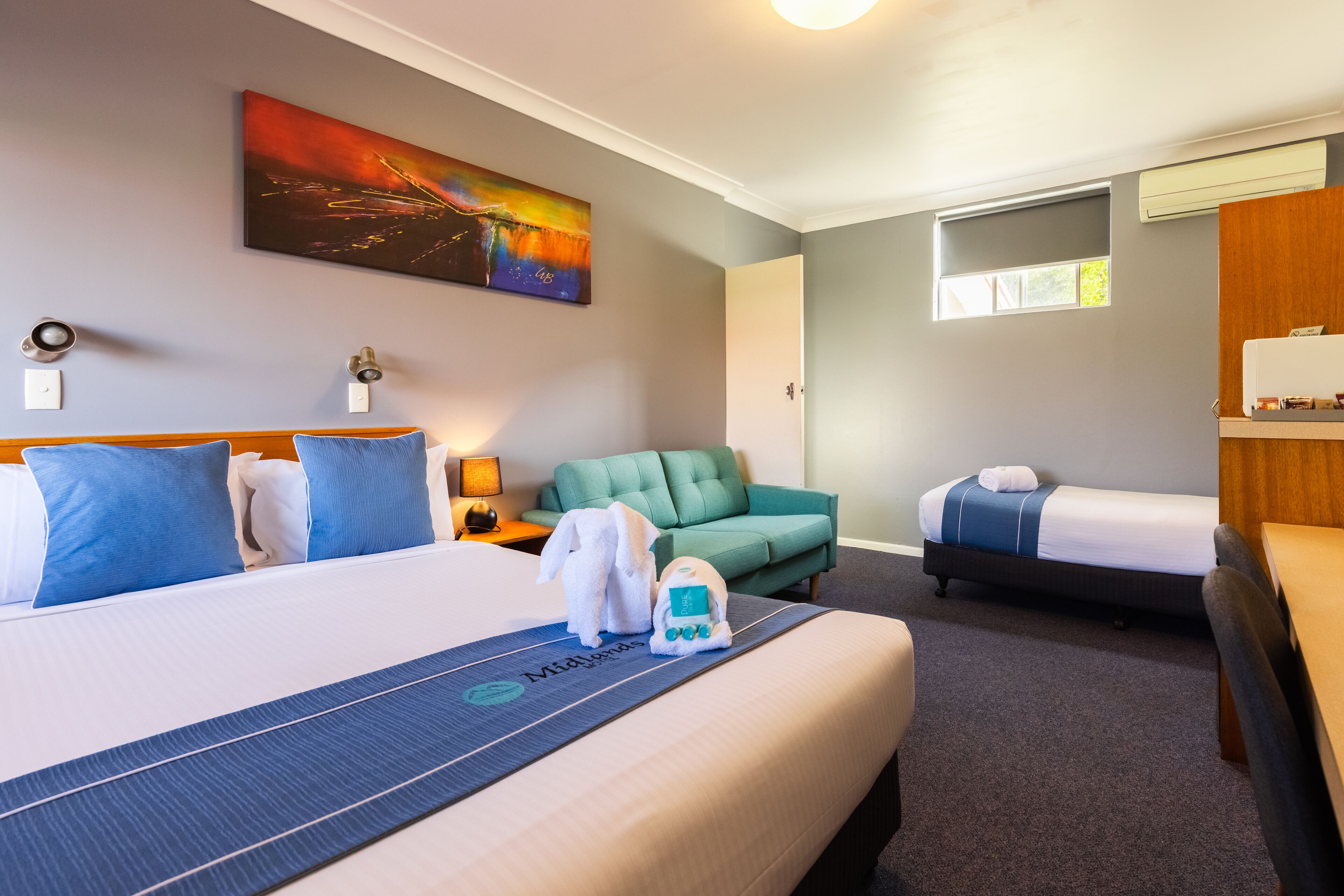 Ein Hotelzimmer mit einem großen Bett, einem grünen Sessel, einem kleineren Bett und einem Schreibtisch mit Stuhl.