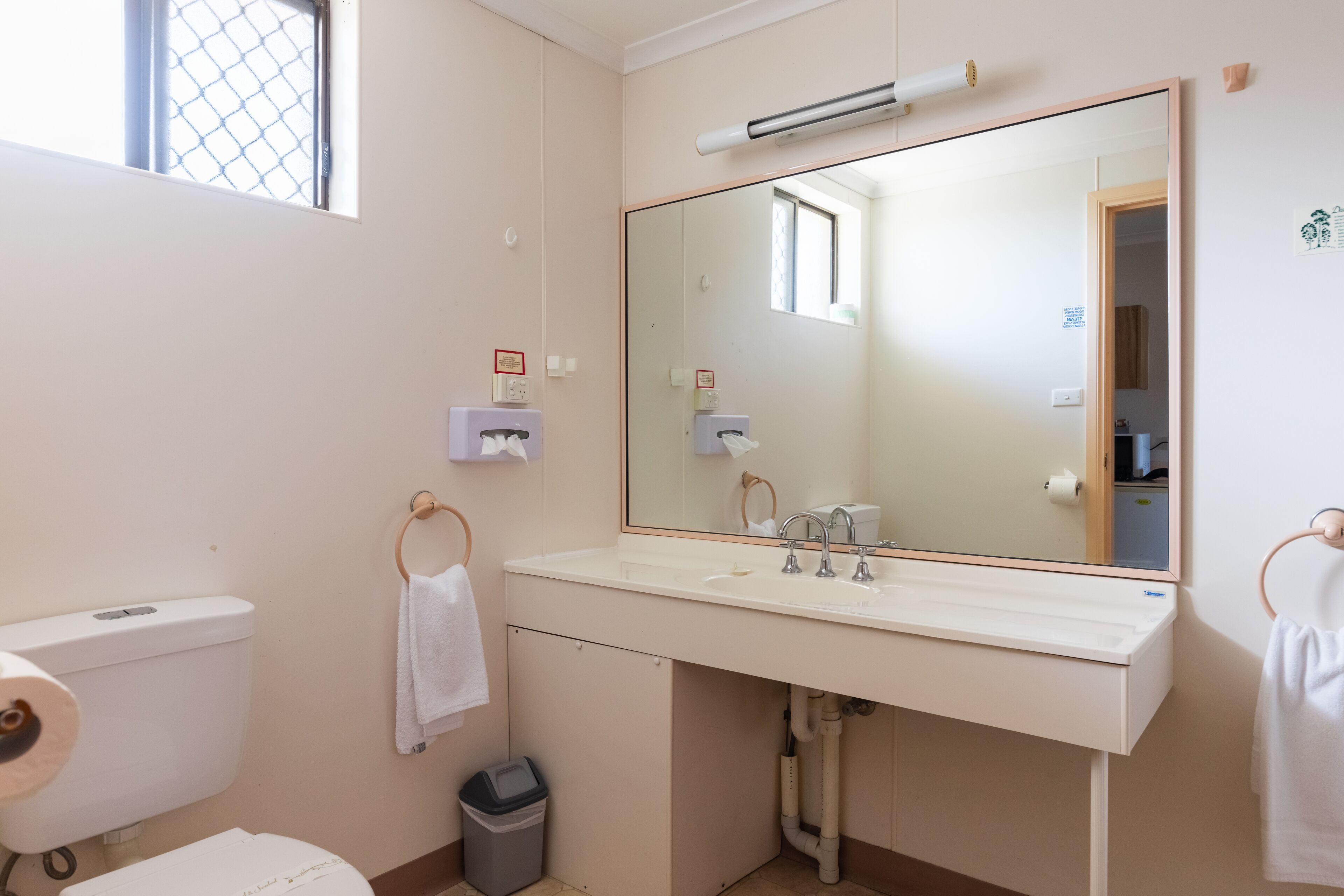 Un bagno con lavandino, specchio e servizi igienici bianchi. È presente un porta asciugamani con un asciugamano bianco, un cestino e una piccola finestra.