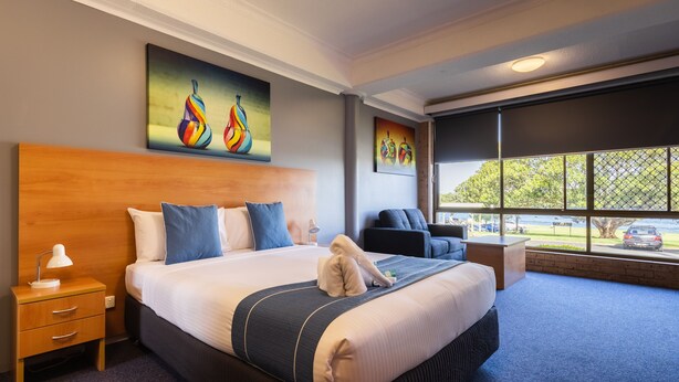 A modern hotel room with a large bed, a wooden headboard, a nightstand, a desk, and a view of the outdoors through large windows.