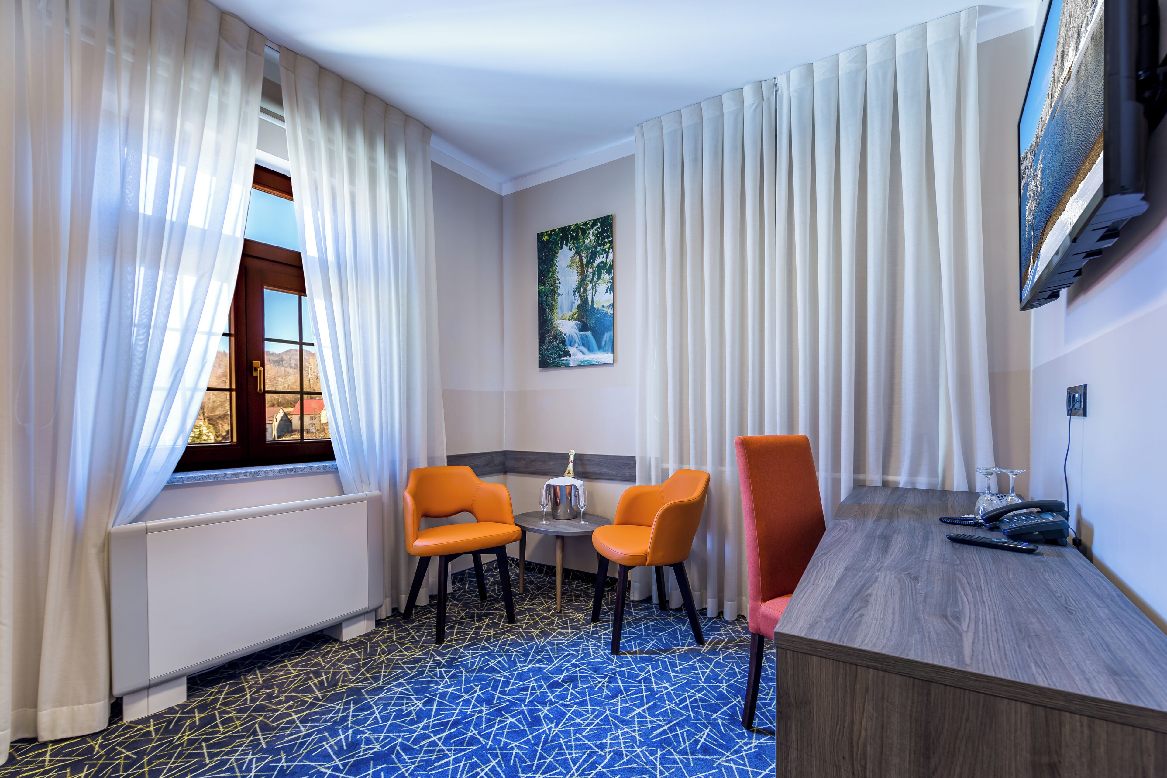 Une chambre d’hôtel avec une grande fenêtre, deux fauteuils orange, une petite table avec un verre, un bureau en bois et un téléviseur fixé au mur.