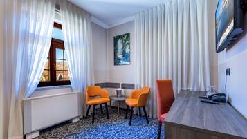 Une chambre d’hôtel avec une grande fenêtre, deux fauteuils orange, une petite table avec un verre, un bureau en bois et un téléviseur fixé au mur.