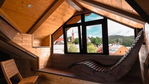 Interior de madera con techo inclinado, un banco y vista a una iglesia y edificios.