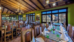 Un comedor con mesas y sillas de madera, ventanas amplias y una araña.