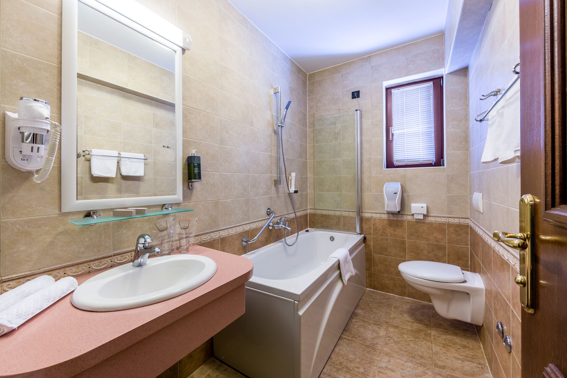 A bathroom with a sink, bathtub, and toilet. There is a mirror, a glass shelf, and a window with blinds.