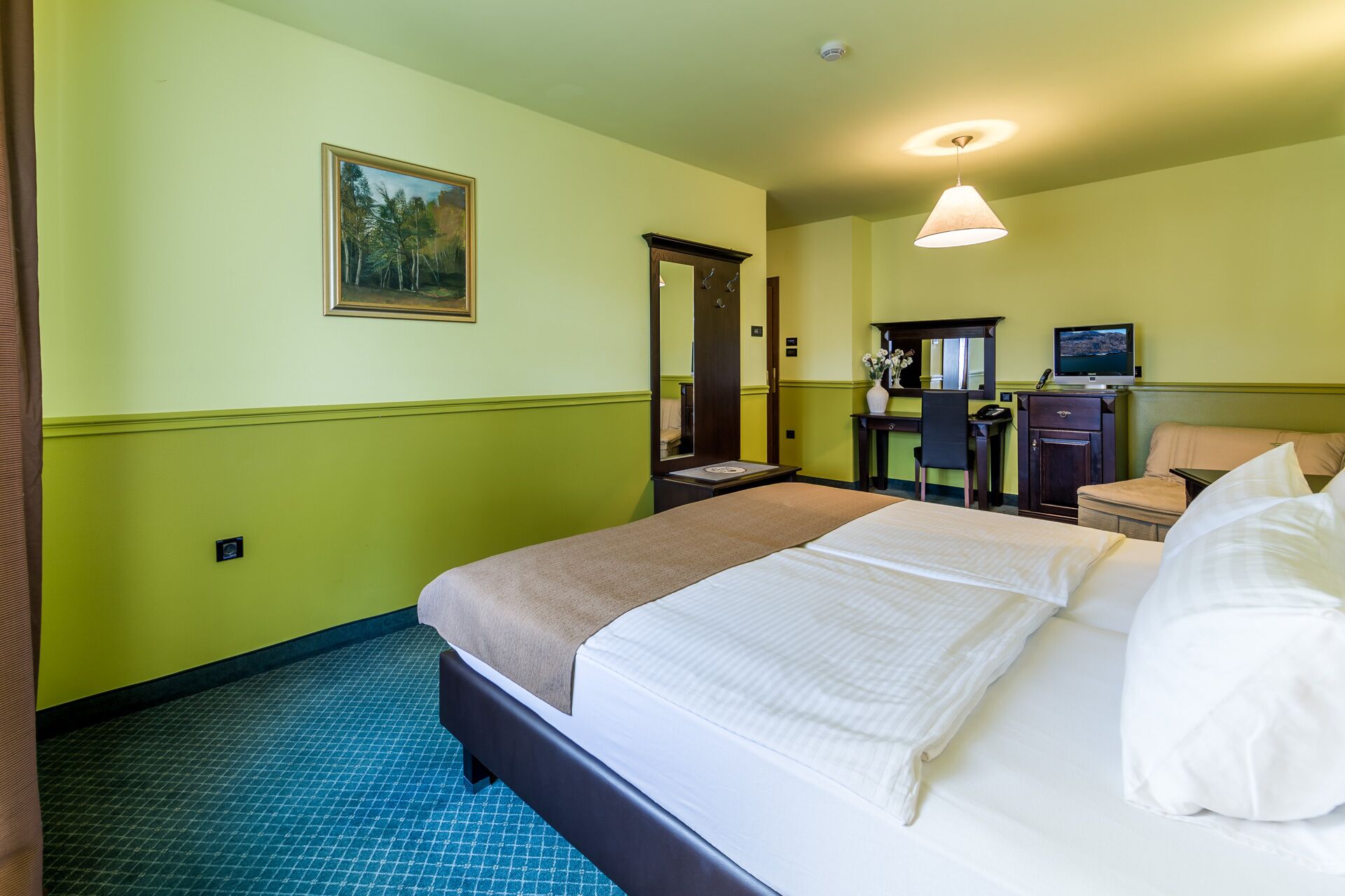 Una habitación de hotel con cama, escritorio, silla, espejo y un cuadro en la pared.