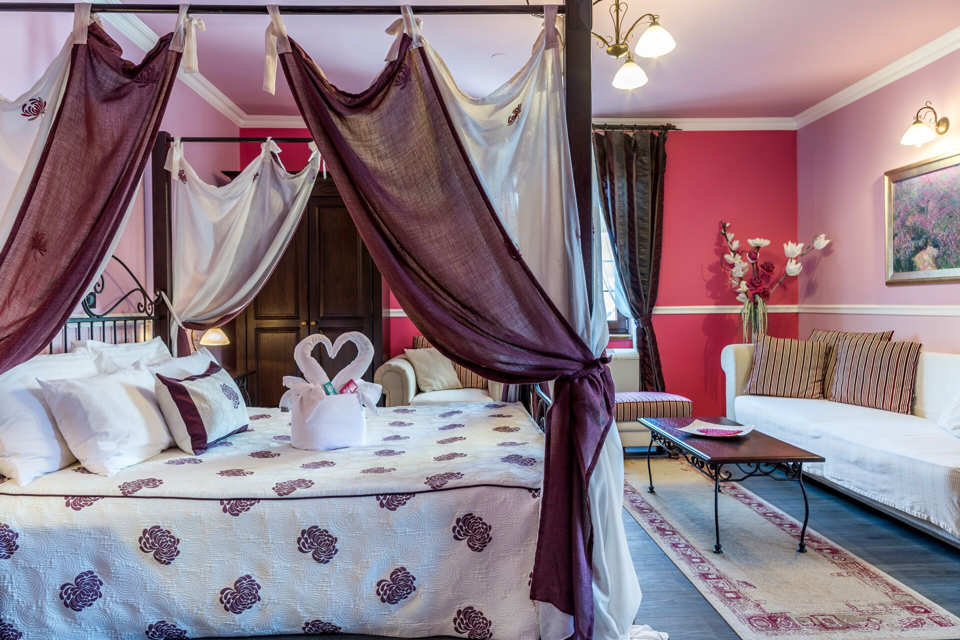 A four-poster canopy bed with a floral bedspread, a bedside table with a vase of flowers, and a sofa with striped cushions.