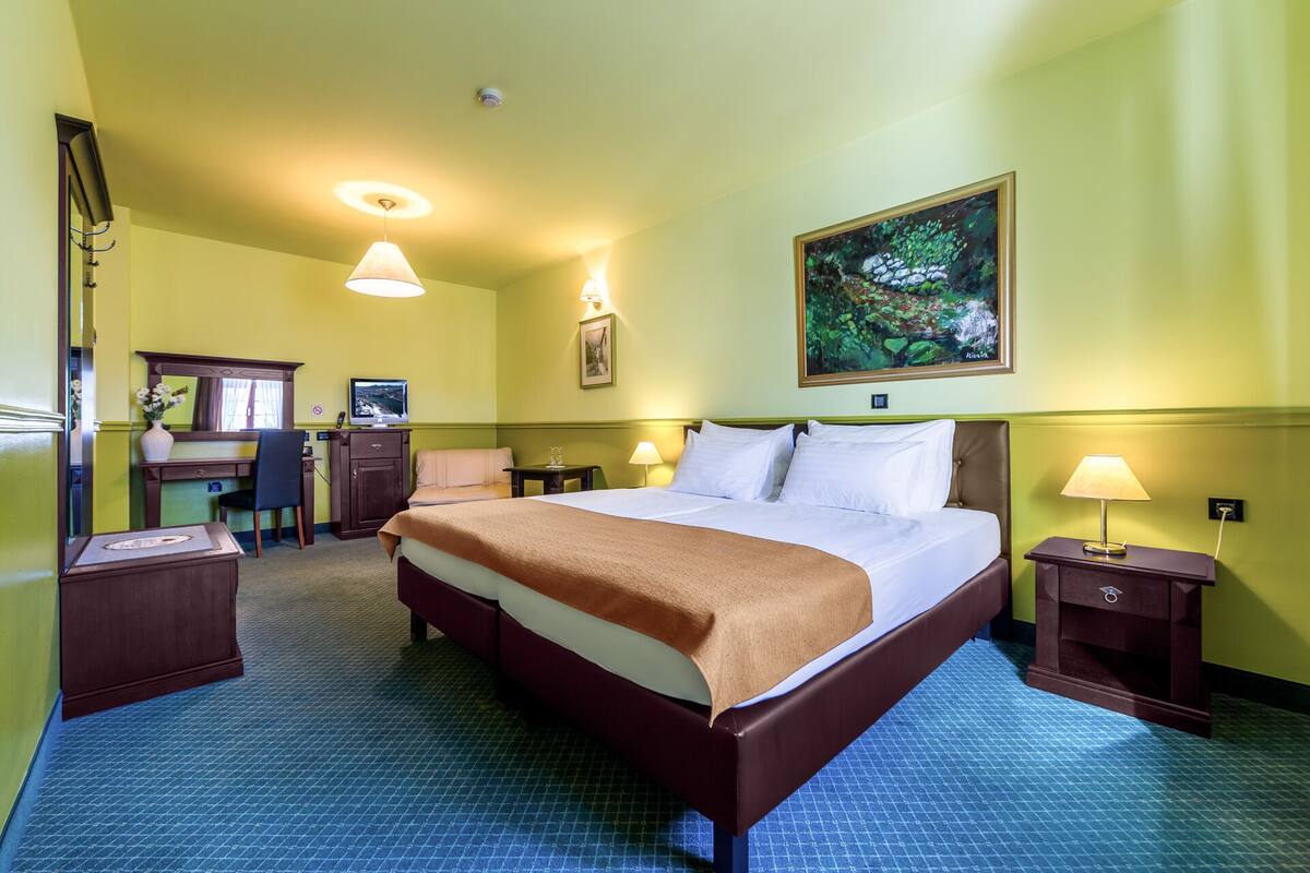 A hotel room with a large bed, a desk with a chair, a painting on the wall, and a desk lamp.