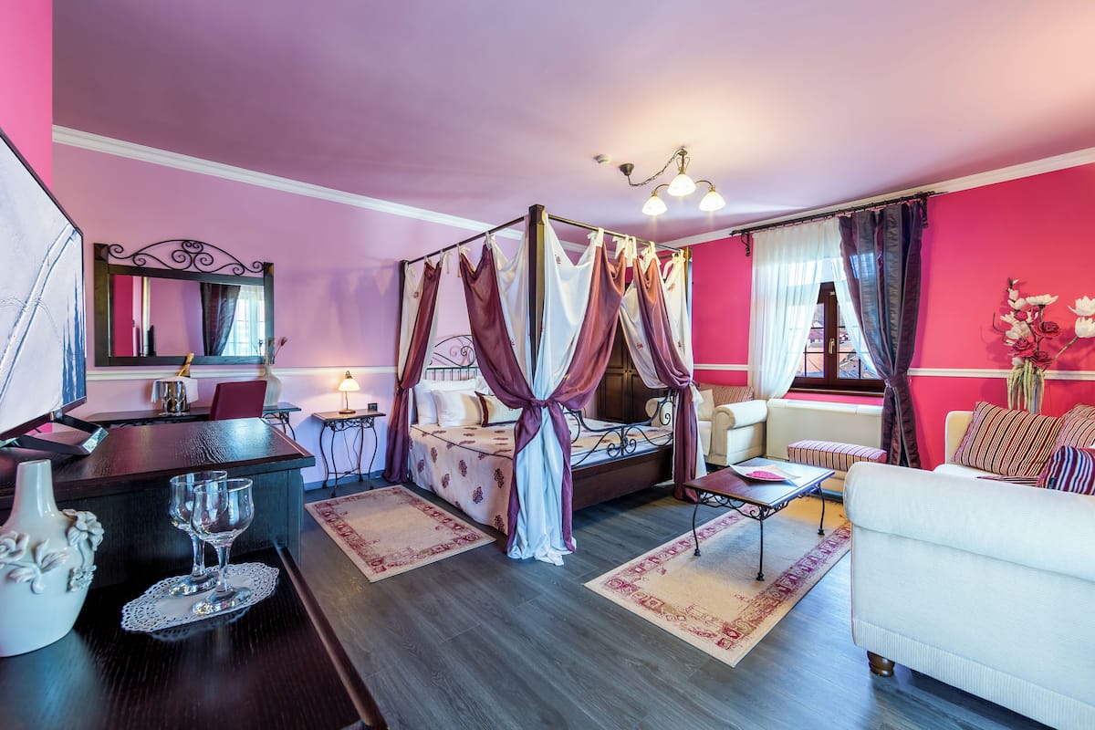 A room with a four-poster bed, a sitting area with a sofa and armchairs, a dining table with chairs, and a small table with two wine glasses.