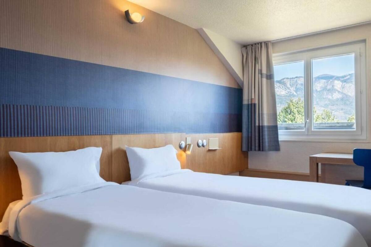 Habitación de hotel con dos camas, cabecero de madera, una mesita y vistas a montañas a través de una ventana.