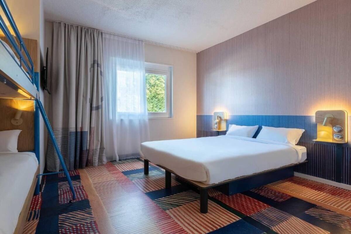 Una habitación de hotel con cama, ventana con cortinas, una escalera y luces fijadas a la pared.