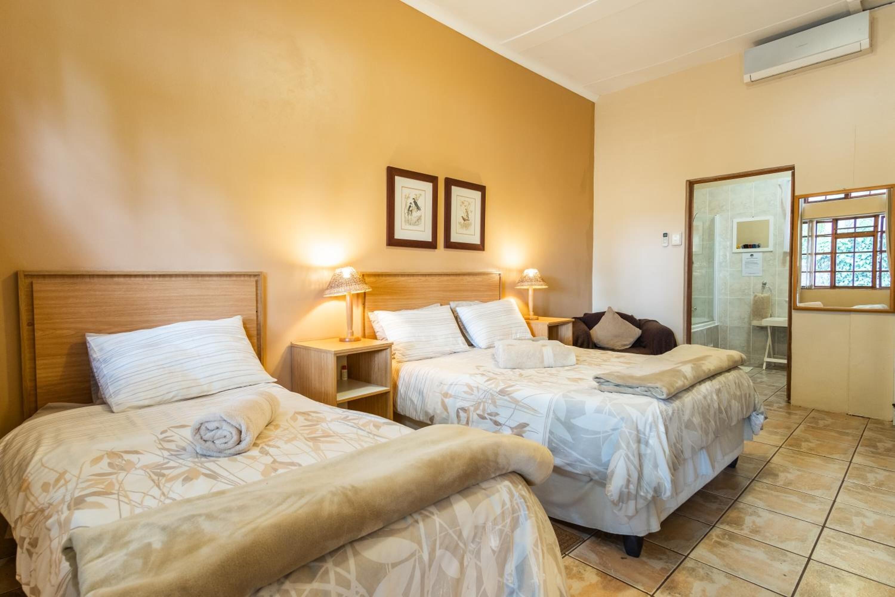 Quarto de hotel com duas camas, cada uma com cabeceira, abajures e uma toalha enrolada em uma delas. Há quadros nas paredes acima das camas e uma janela com vista para o verde.