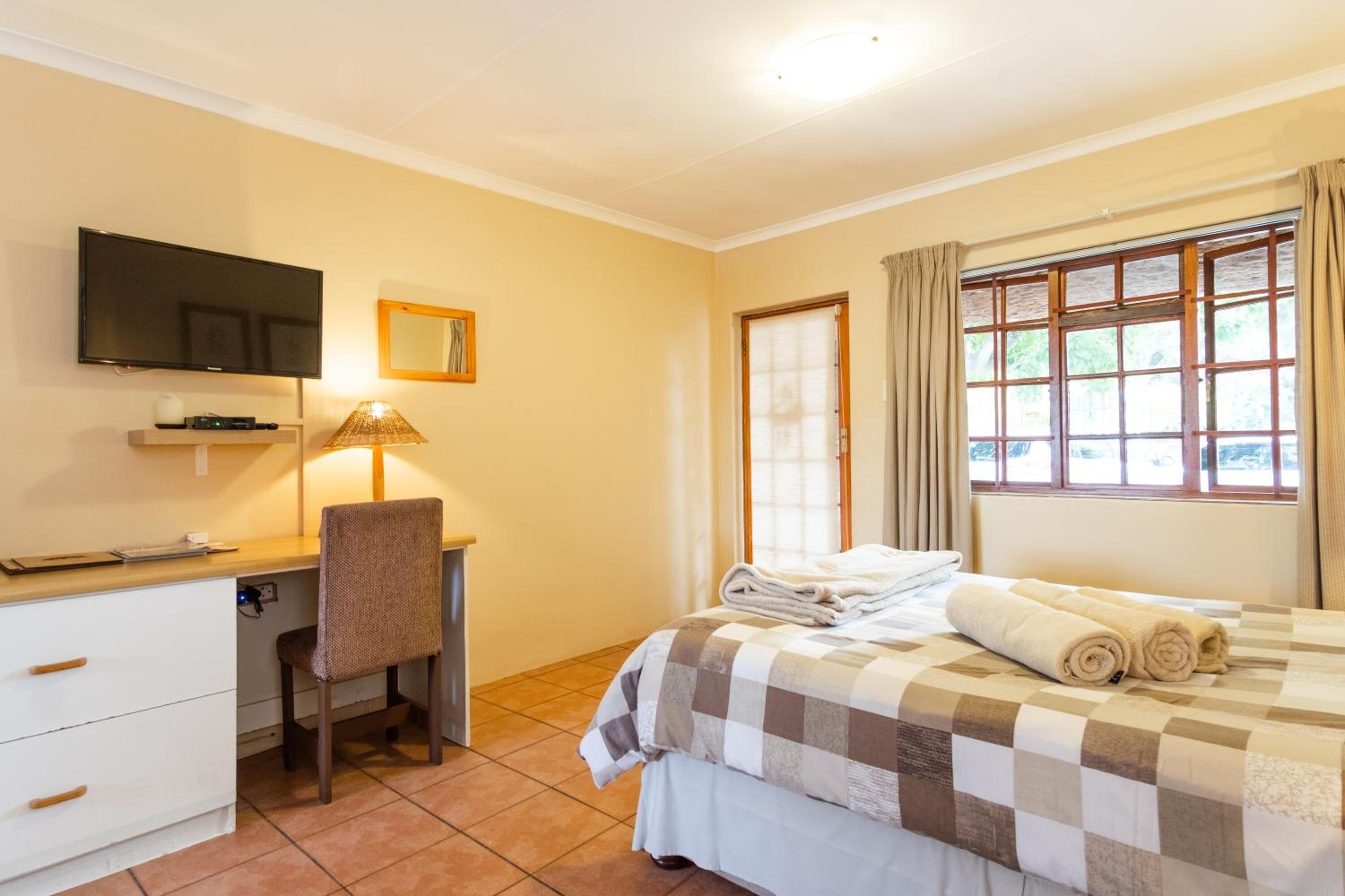 Una habitación de hotel con cama, escritorio, silla, televisor y ventana con cortinas.