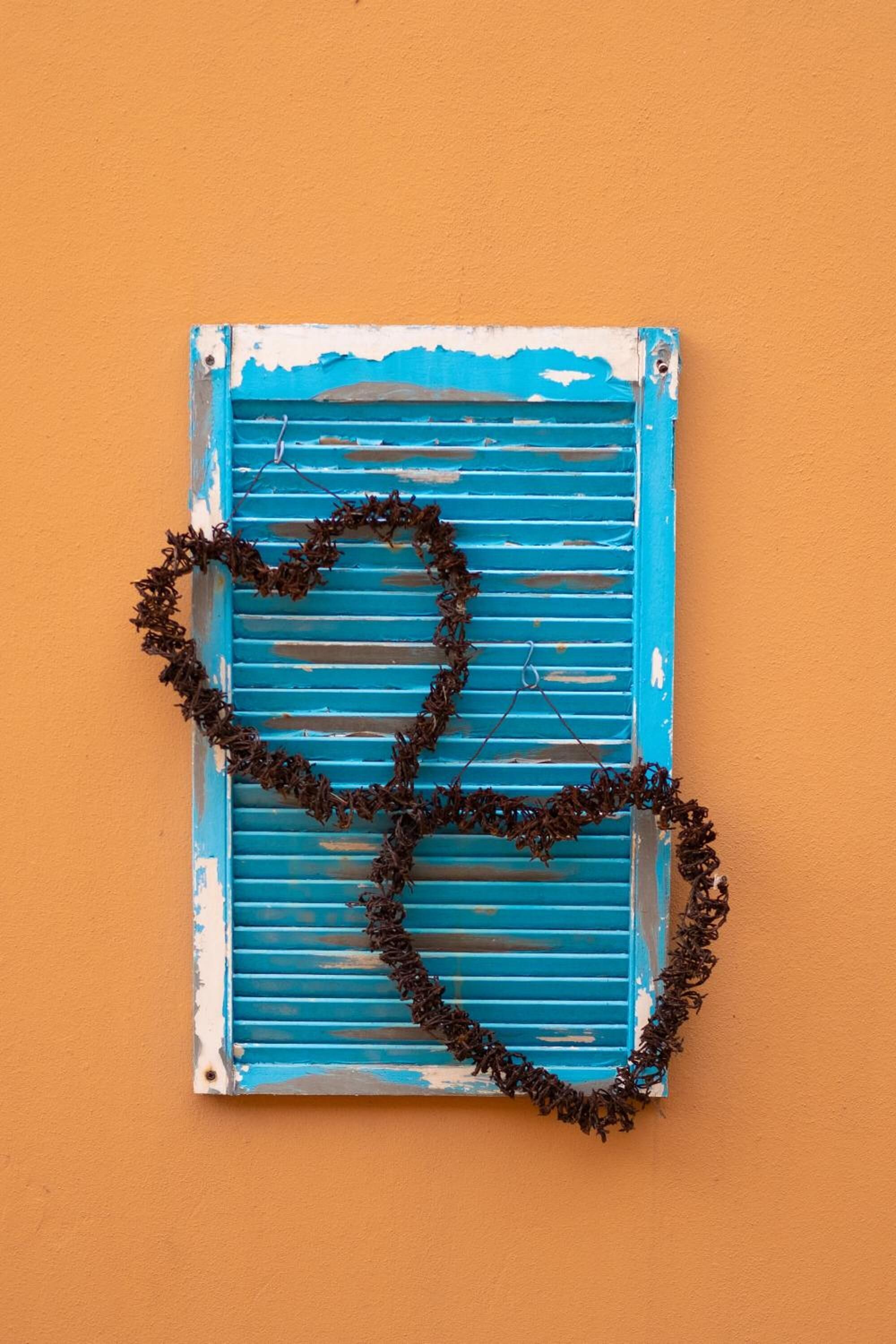 A blue shutter with a heart-shaped decoration made of dried plants against an orange wall.