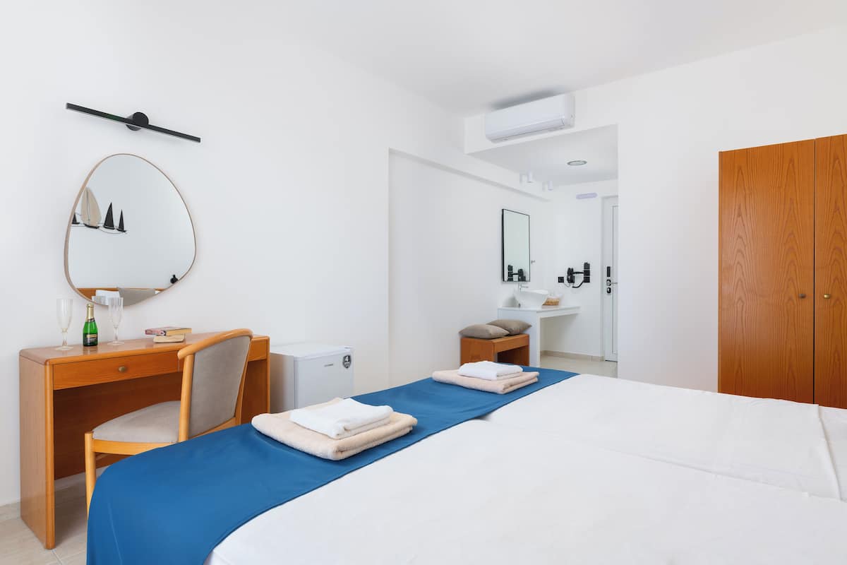 A hotel room with a bed, a desk, a chair, a mirror, a wardrobe, and a bathroom with a sink and mirror.