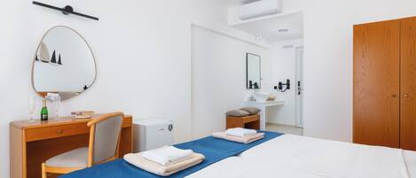 A hotel room with a bed, a desk, a chair, a mirror, a wardrobe, and a bathroom with a sink and mirror.