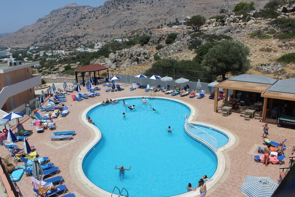 A swimming pool with lounge chairs and umbrellas, surrounded by buildings and a mountainous landscape.