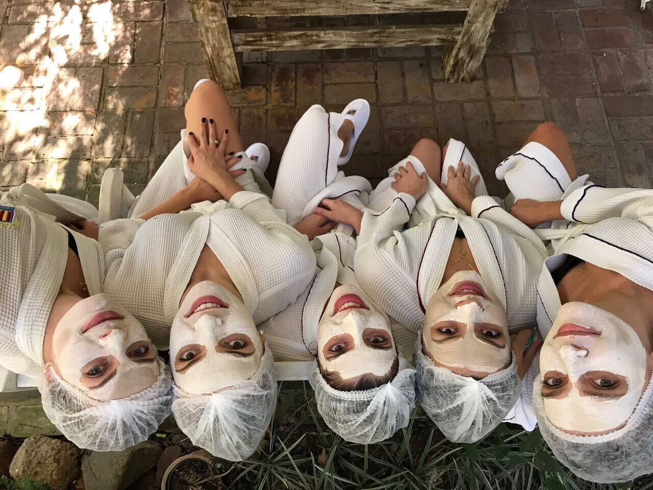 A group of people lying on a bench outdoors, wearing white robes and slippers, with a brick wall and greenery in the background.