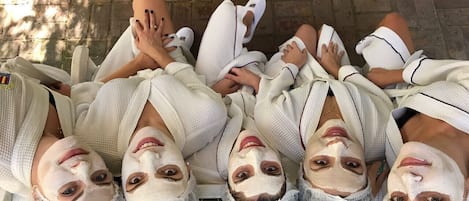 A group of people lying on a bench outdoors, wearing white robes and slippers, with a brick wall and greenery in the background.