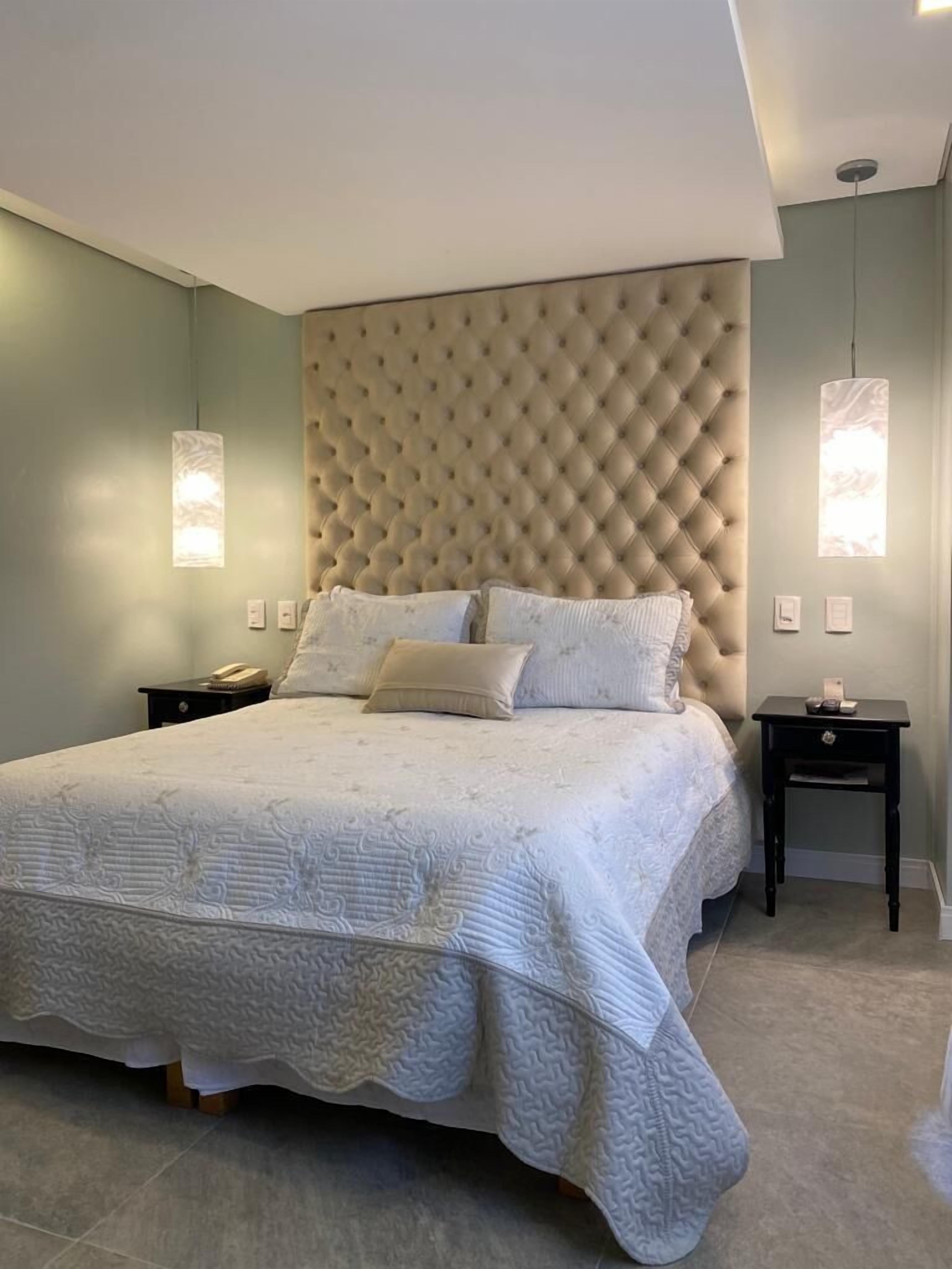 Un dormitorio con cabecera capitonada, una cama grande con colcha estampada, dos mesitas de noche con lámparas y una paleta de colores neutros.