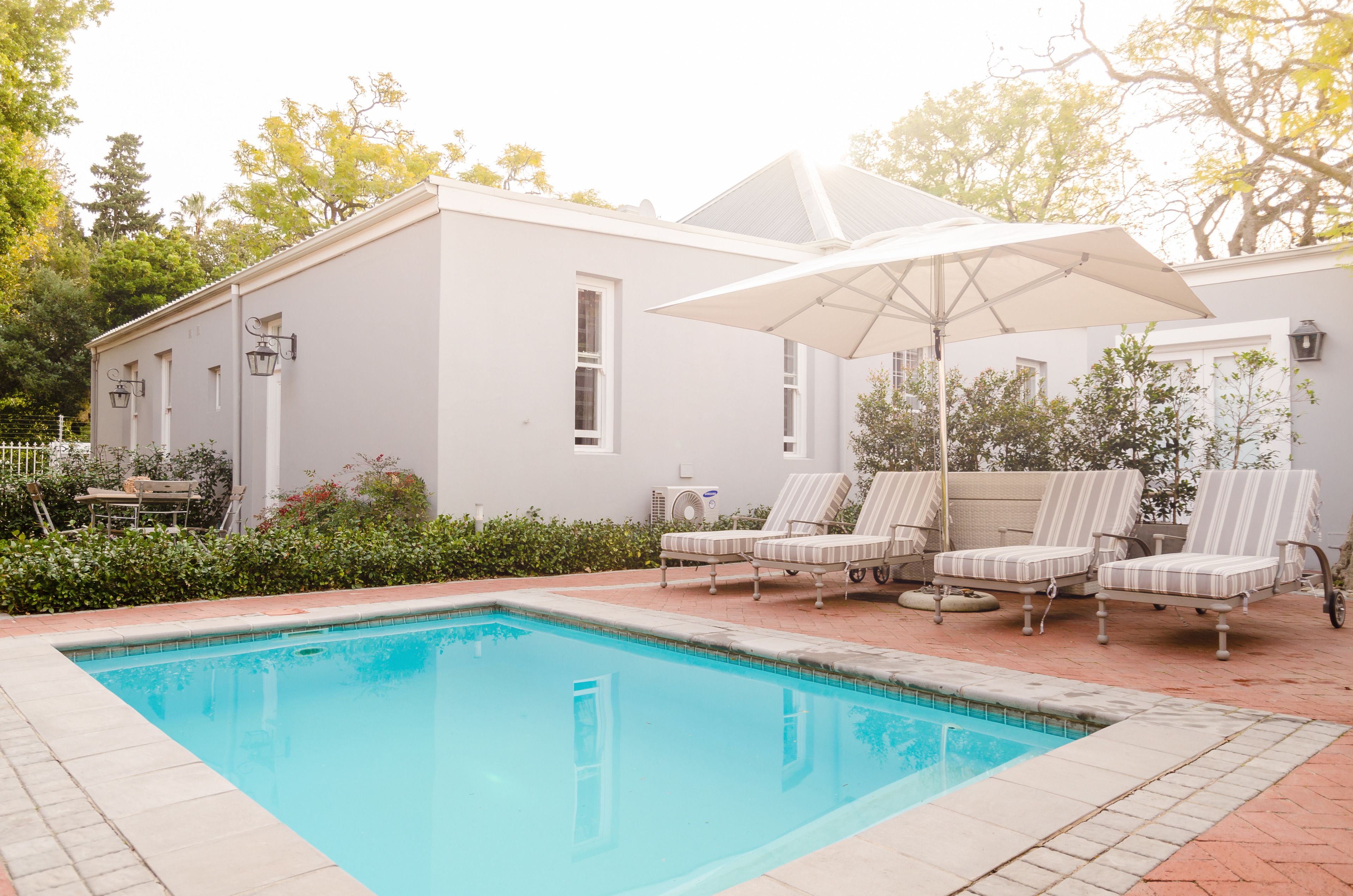 A swimming pool with lounge chairs and an umbrella, surrounded by a white building with windows and a brick pathway.