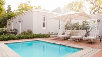 A swimming pool with lounge chairs and an umbrella, surrounded by a white building with windows and a brick pathway.